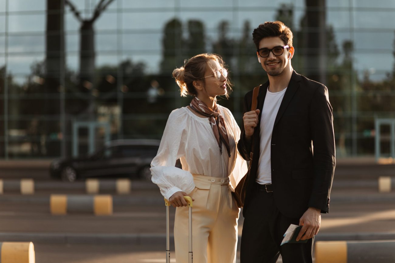 Brunette man in black suit and sunglasses smiles. Attractive blonde woman in white blouse looks at