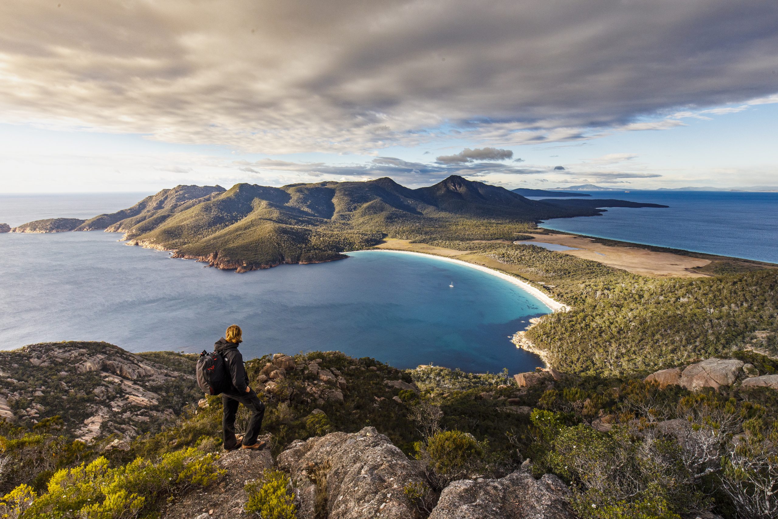 Young male hiker with backpack standing on top of mountain overlooking coast at wineglass bay